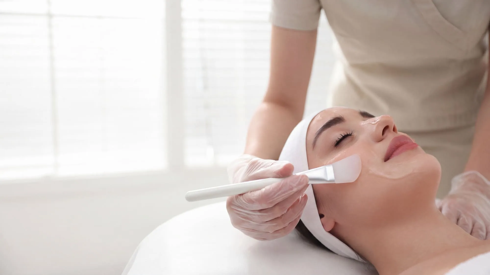 Wide view of a medical-grade facial in progress, a gloved clinician brushing treatment onto a reclining patient's skin in a bright, daylit treatment room