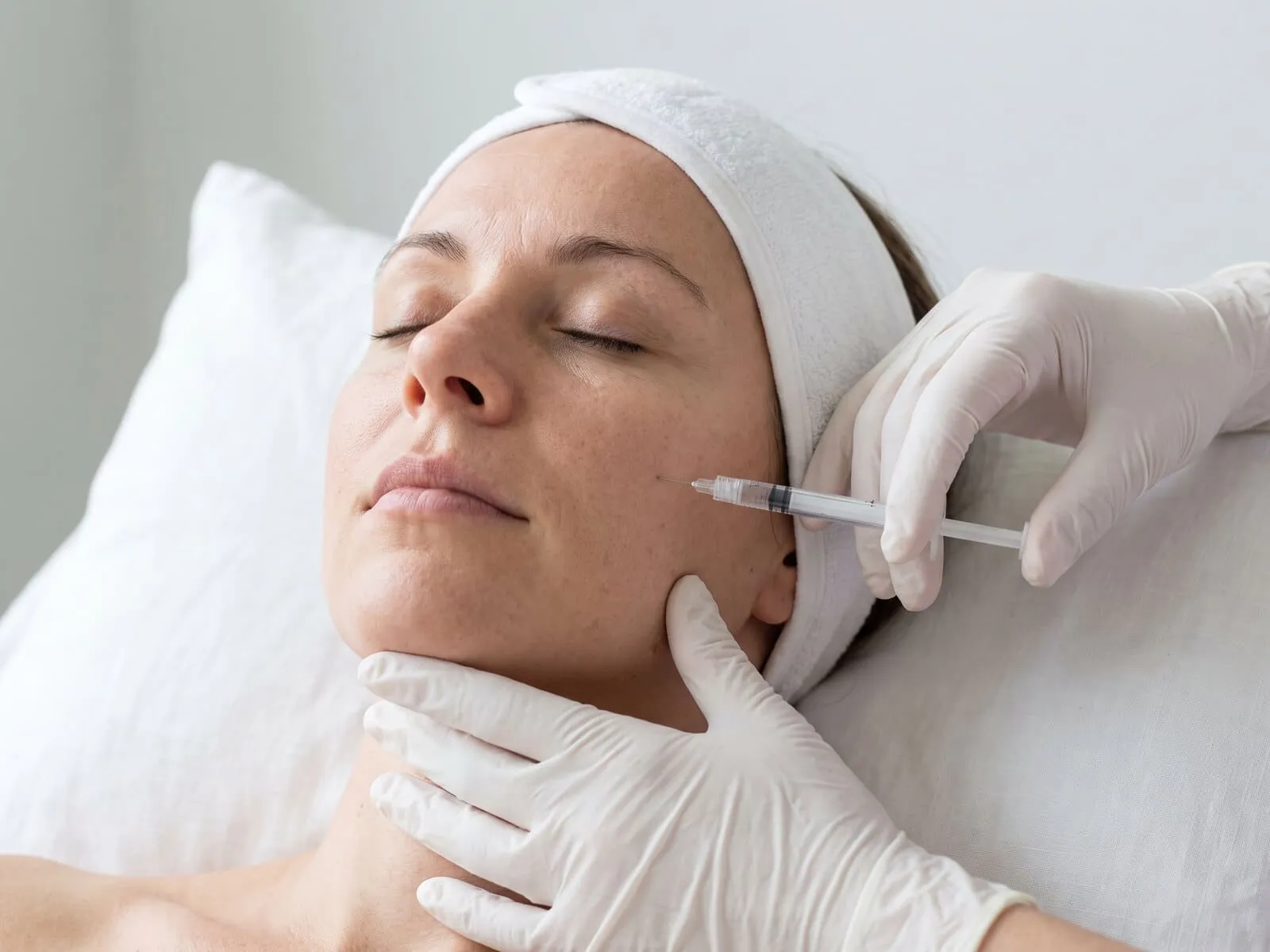 Close view of a gloved clinician placing a fine dermal filler needle at the mid-cheek of a reclining patient