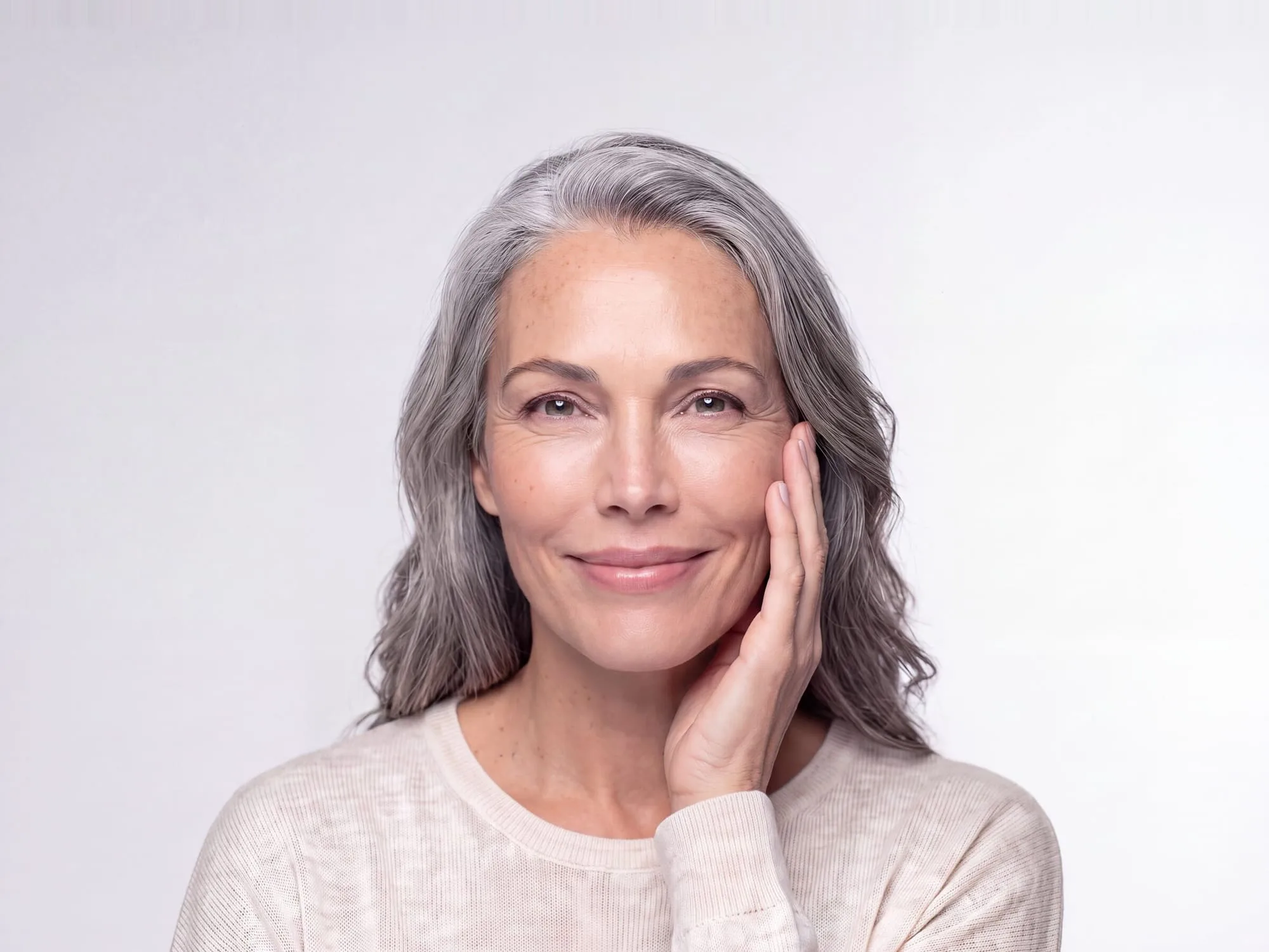 Close-up of a woman in her fifties with long silver-grey hair resting her fingertips lightly against her cheek, smiling gently to camera in soft studio light against a plain pale grey background