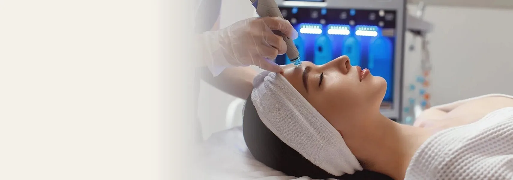 A HydraFacial in progress: a gloved practitioner guiding the vortex handpiece across a reclining patient's cheek, with the device and its serum bottles behind them