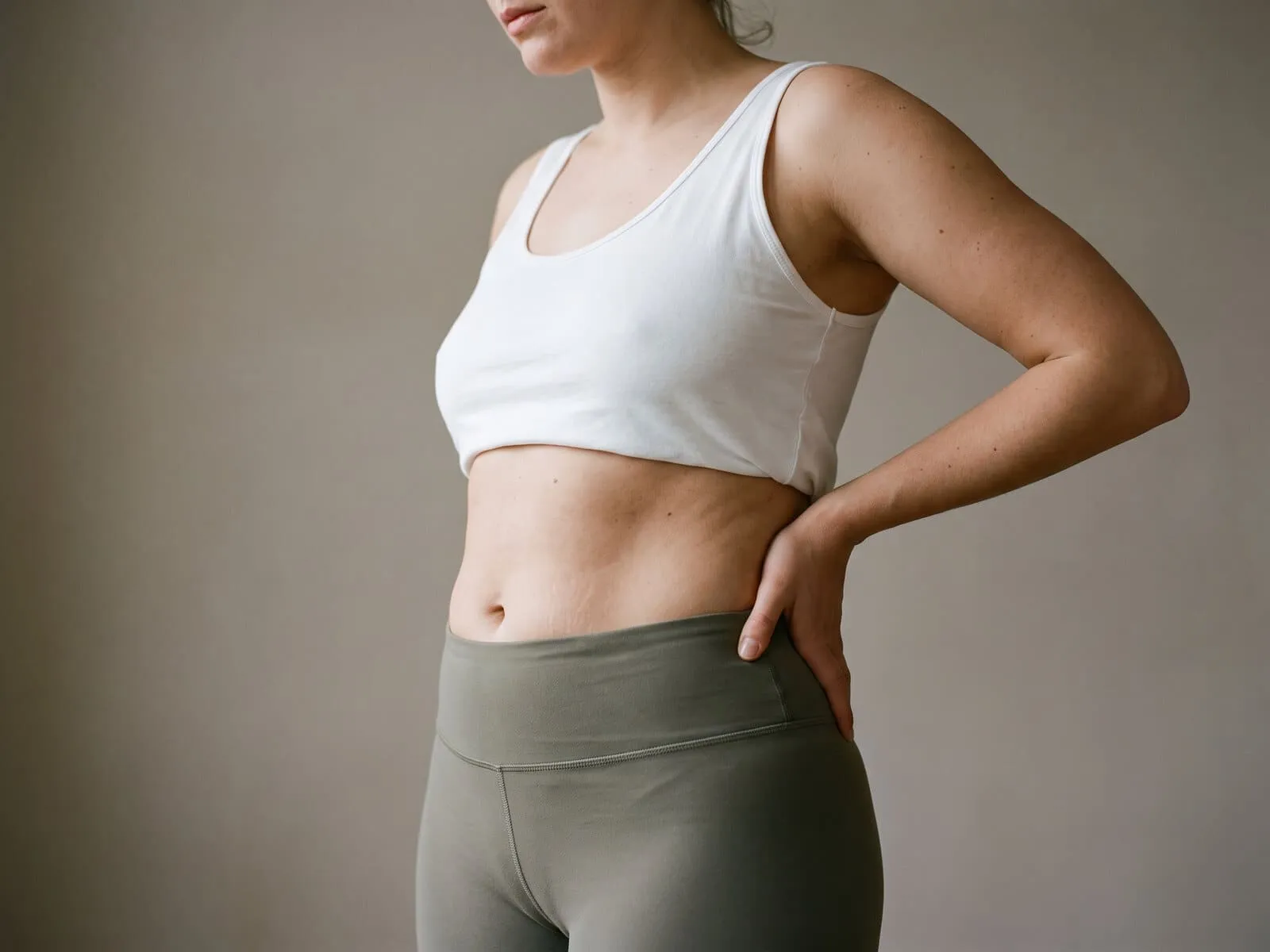 A woman standing with a hand on her waist in a cropped white top and olive leggings, showing the untouched midsection and waist clients ask about when tone rather than weight is the goal