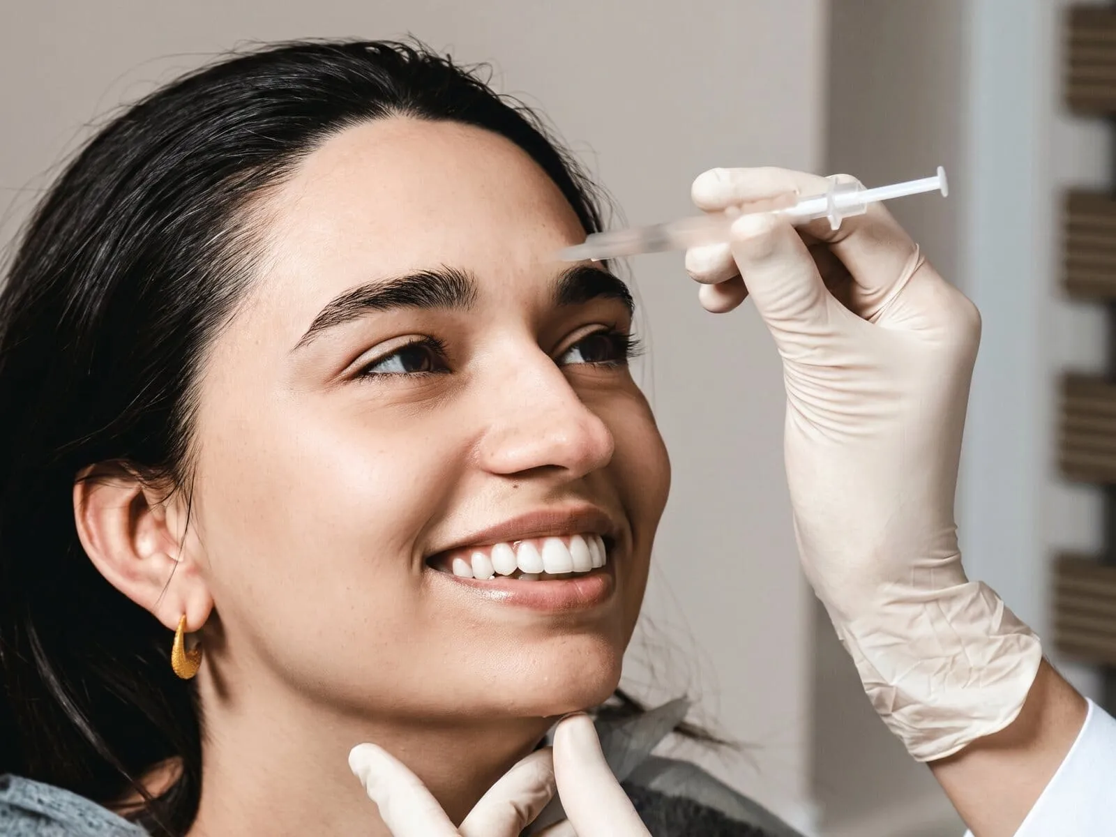 A seated patient smiling while a gloved clinician holds a fine-gauge syringe at her outer brow, the area neurotoxins treat
